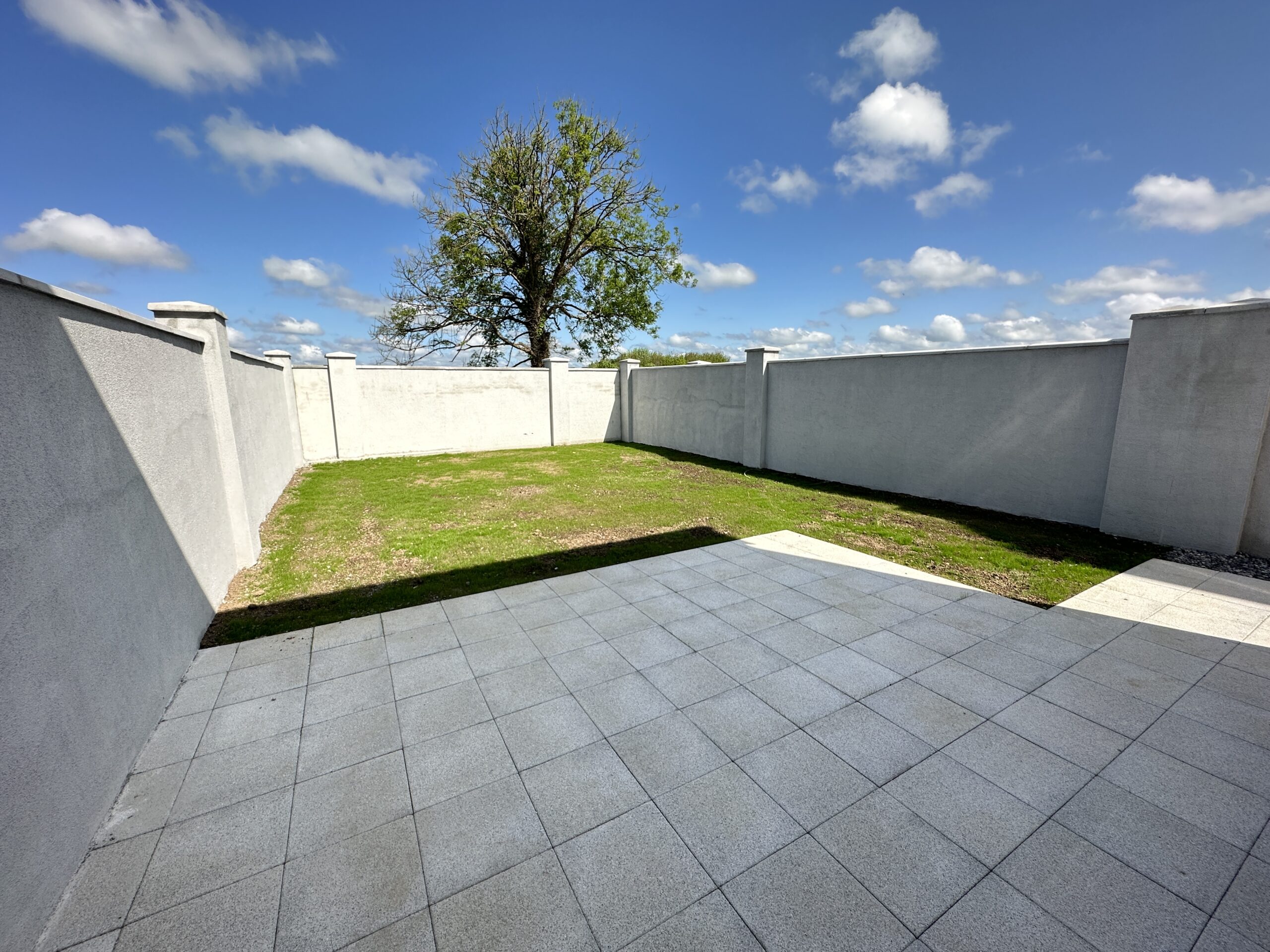 A minimalist outdoor space with gray paved tiles leading to a small grassy area, encircled by high white walls under a blue sky with fluffy clouds, perfect for a home page background.